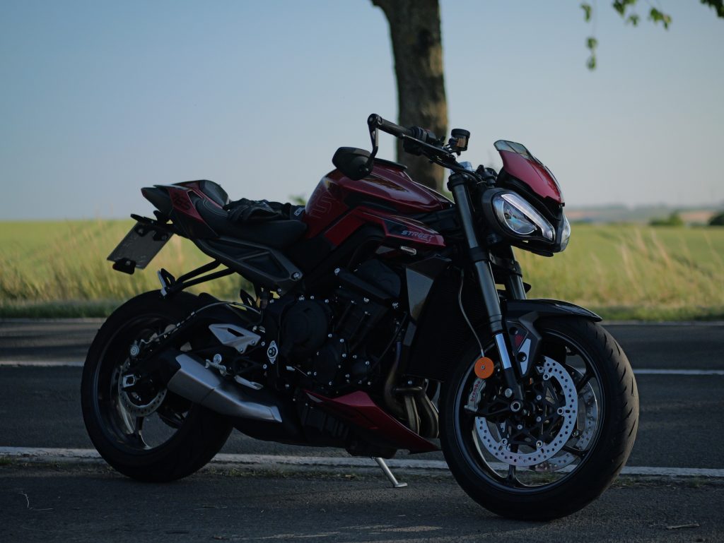 Motorrad - Triumph Street Triple 765RS mit eingeschalteter Beleuchtung steht an einer Landstraße mit einem Feld im Hintergrund