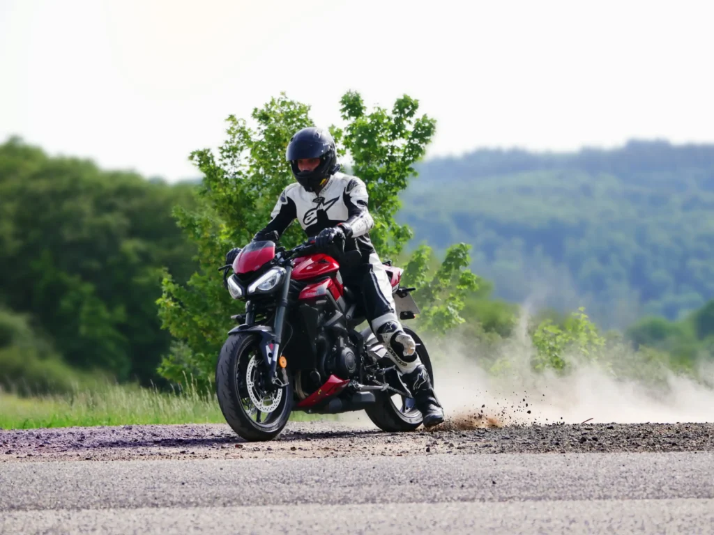 Motorradfahrer in weißer Lederkombi fährt auf einem Naked Bike (Triumph Street Triple 765 RS) mit durchdrehendem Reifen auf Schotter los. Das linke Bein streift dabei noch leicht über den Boden. Hinter ihm wirbelt eine Staubwolke auf und Steine fliegen hoch.