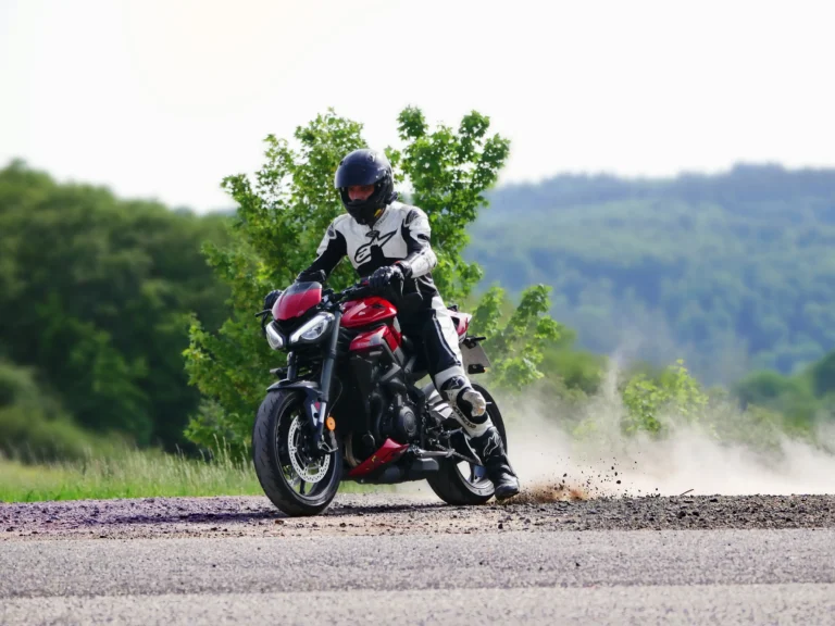 Motorradfahrer in weißer Lederkombi fährt auf einem Naked Bike (Triumph Street Triple 765 RS) mit durchdrehendem Reifen auf Schotter los. Das linke Bein streift dabei noch leicht über den Boden. Hinter ihm wirbelt eine Staubwolke auf und Steine fliegen hoch.