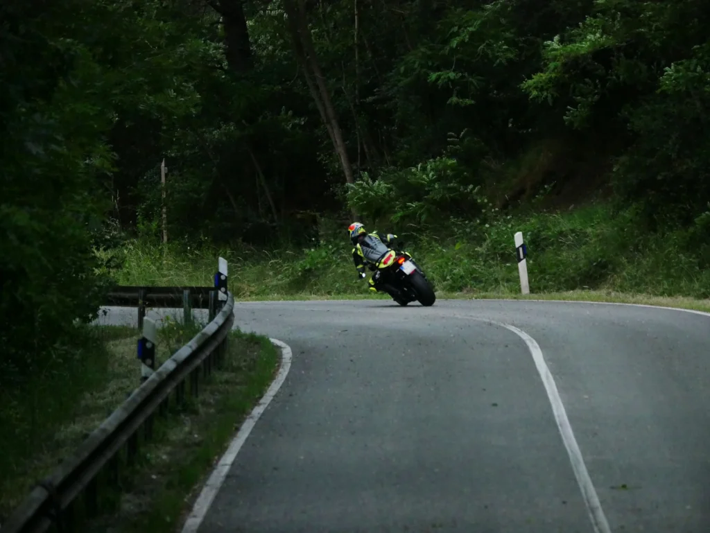 Motorradfahrer von hinten beim Bremsen in Schräglage in einer engen Linkskurve auf einer Landstraße. Die Straße führt durch einen Wald. Der Hintergrund ist von Bäumen und Pflanzen gesäumt. Am linken Rand sieht man eine Leitplanke.Der Motorradfahrer trägt eine Neon-gelbe und graue Lederkombi mit einem passenden bunten Helm. Er fährt ein Naked Bike - Triumph Street Triple 765RS