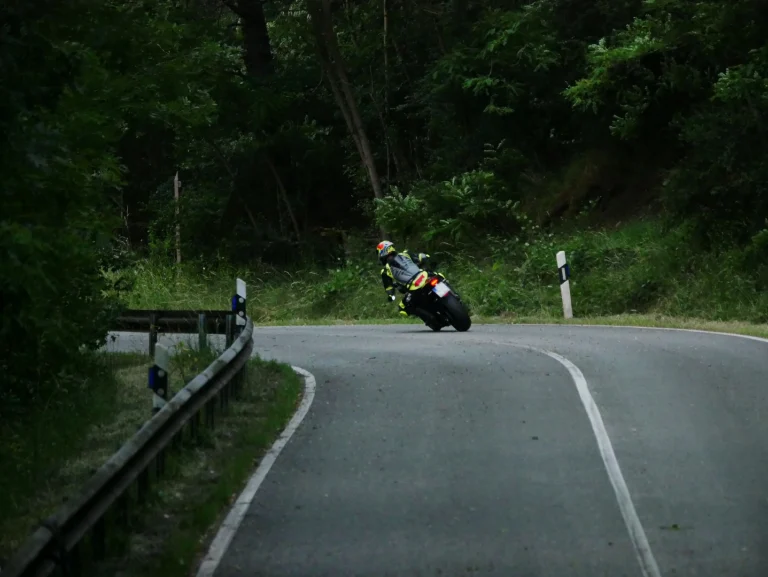 Motorradfahrer von hinten beim Bremsen in Schräglage in einer engen Linkskurve auf einer Landstraße. Die Straße führt durch einen Wald. Der Hintergrund ist von Bäumen und Pflanzen gesäumt. Am linken Rand sieht man eine Leitplanke.Der Motorradfahrer trägt eine Neon-gelbe und graue Lederkombi mit einem passenden bunten Helm. Er fährt ein Naked Bike - Triumph Street Triple 765RS