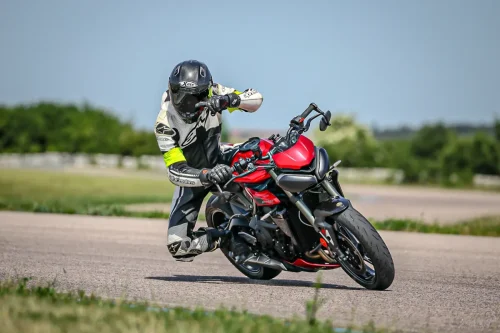 Motorradfahrer mit neon-gelben Armbinden auf der Rennstrecke mit einer Triumph Street Triple 765RS. Er fährt Schräglage im Hanging-Off und schaut Richtung Kurvenausgang und zeigt mit einer Hand dorthin