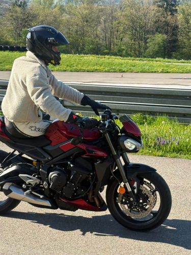 Motorradfahrer fährt seitlich sitzend mit beiden Beinen auf einer Seite des Motorrades und nur einer Hand am Lenker. Der Fahrer trägt einen hellen, dicken Pullover über einer Lederkombi